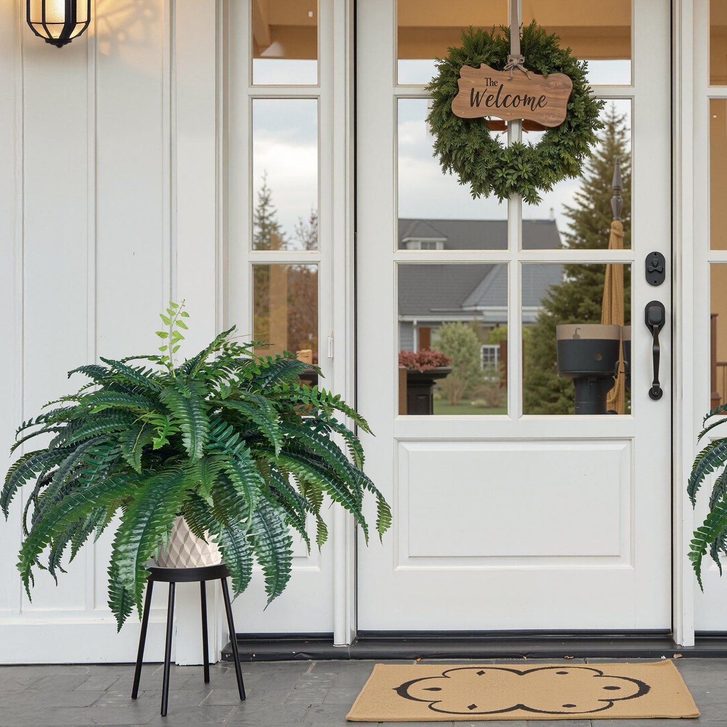 Artificial Boston fern plant in white pot on black stand placed near white front door with welcome wreath