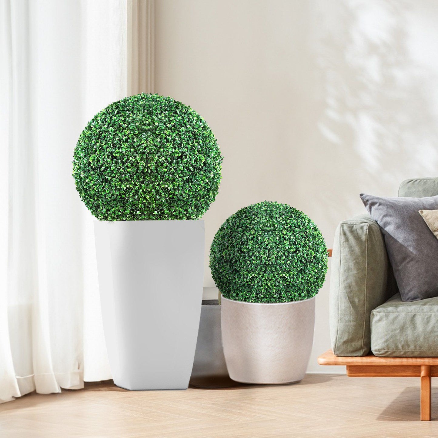 Artificial boxwood topiary balls in white and beige pots displayed in a modern living room next to a gray sofa