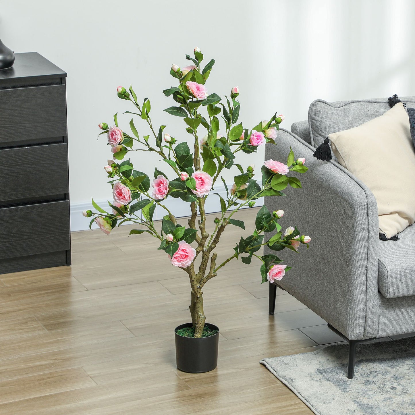 Artificial camellia flowers in pots with pink blooms and green leaves next to gray sofa and black drawer
