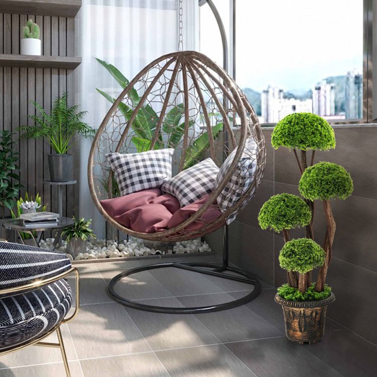 Artificial cedar topiary tree with multiple green rounded foliage in a decorative pot beside a hanging wicker chair with cushions