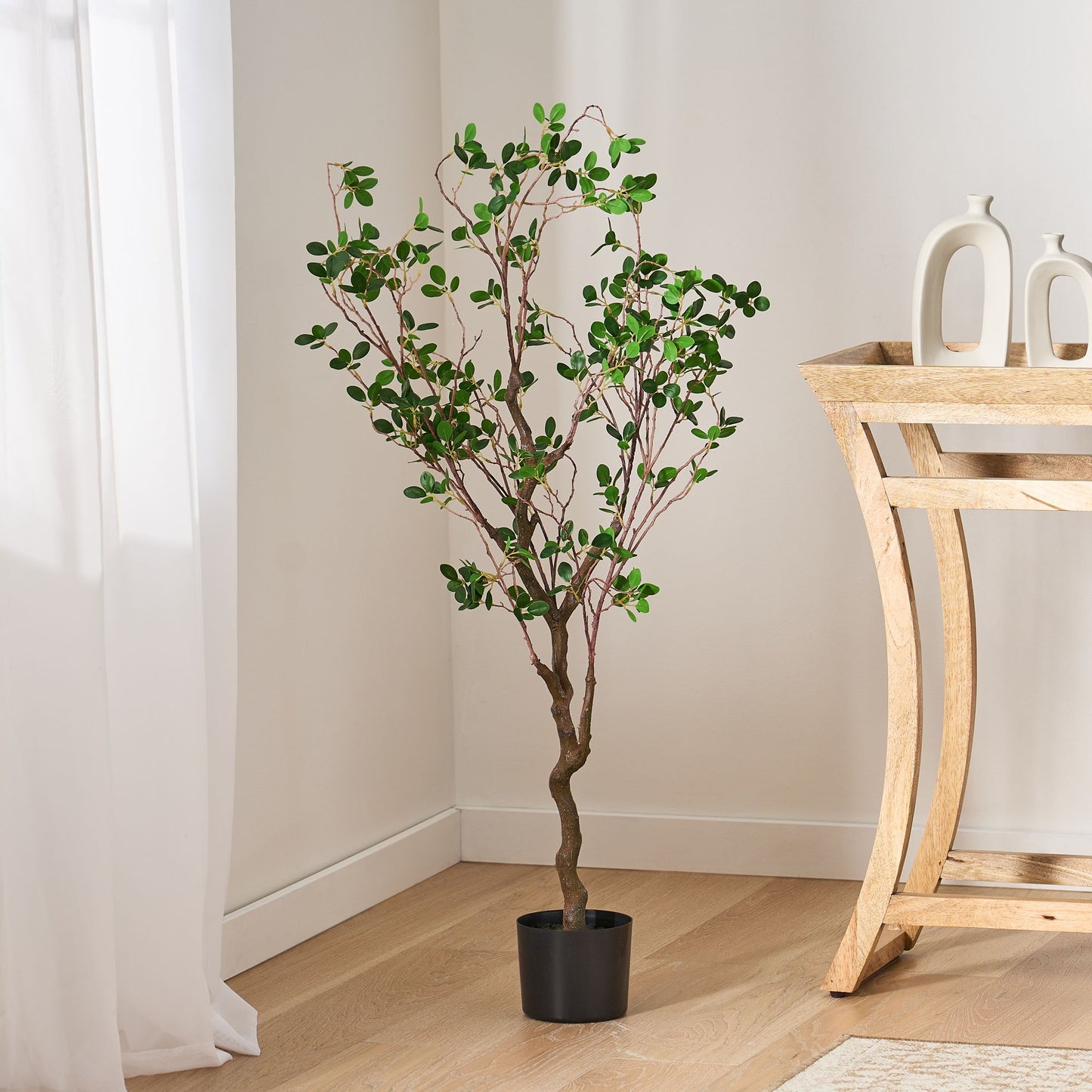 Artificial Milan leaf bonsai tree in black pot placed in modern room corner by wooden table