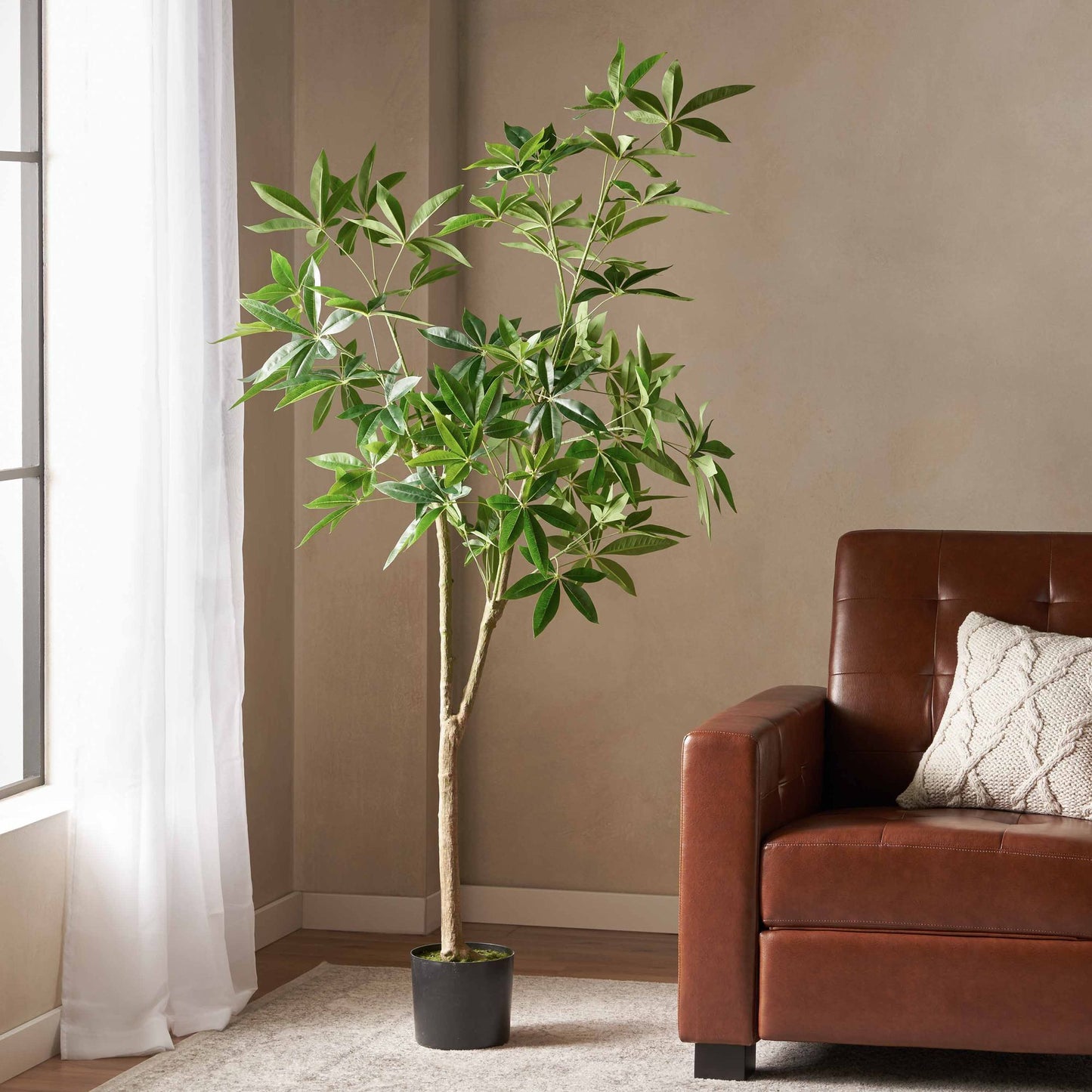 Tall artificial pachira money tree with green leaves in black pot beside brown leather sofa
