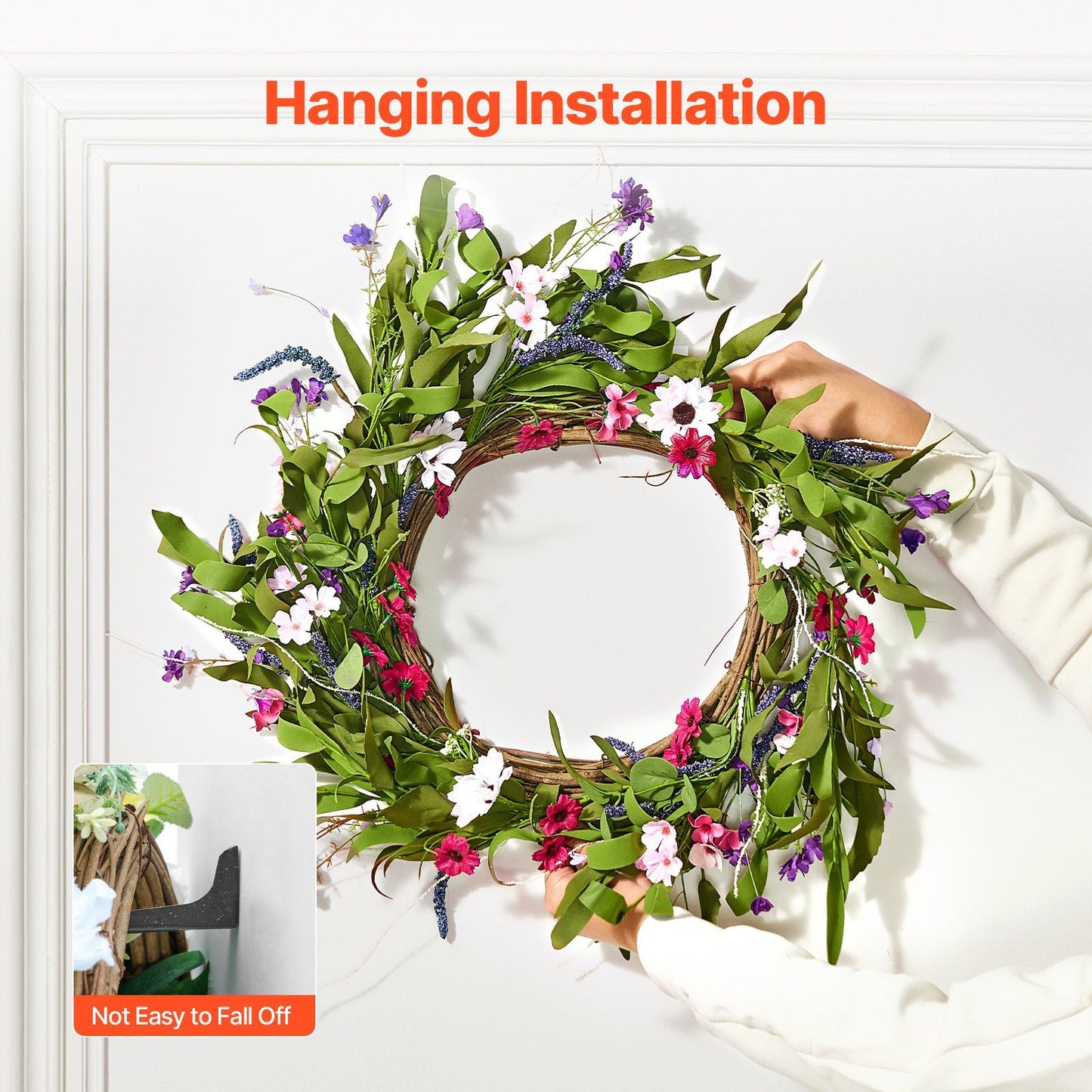 Hands hanging an artificial pink daisy wreath with green leaves and mixed flowers on a white wall