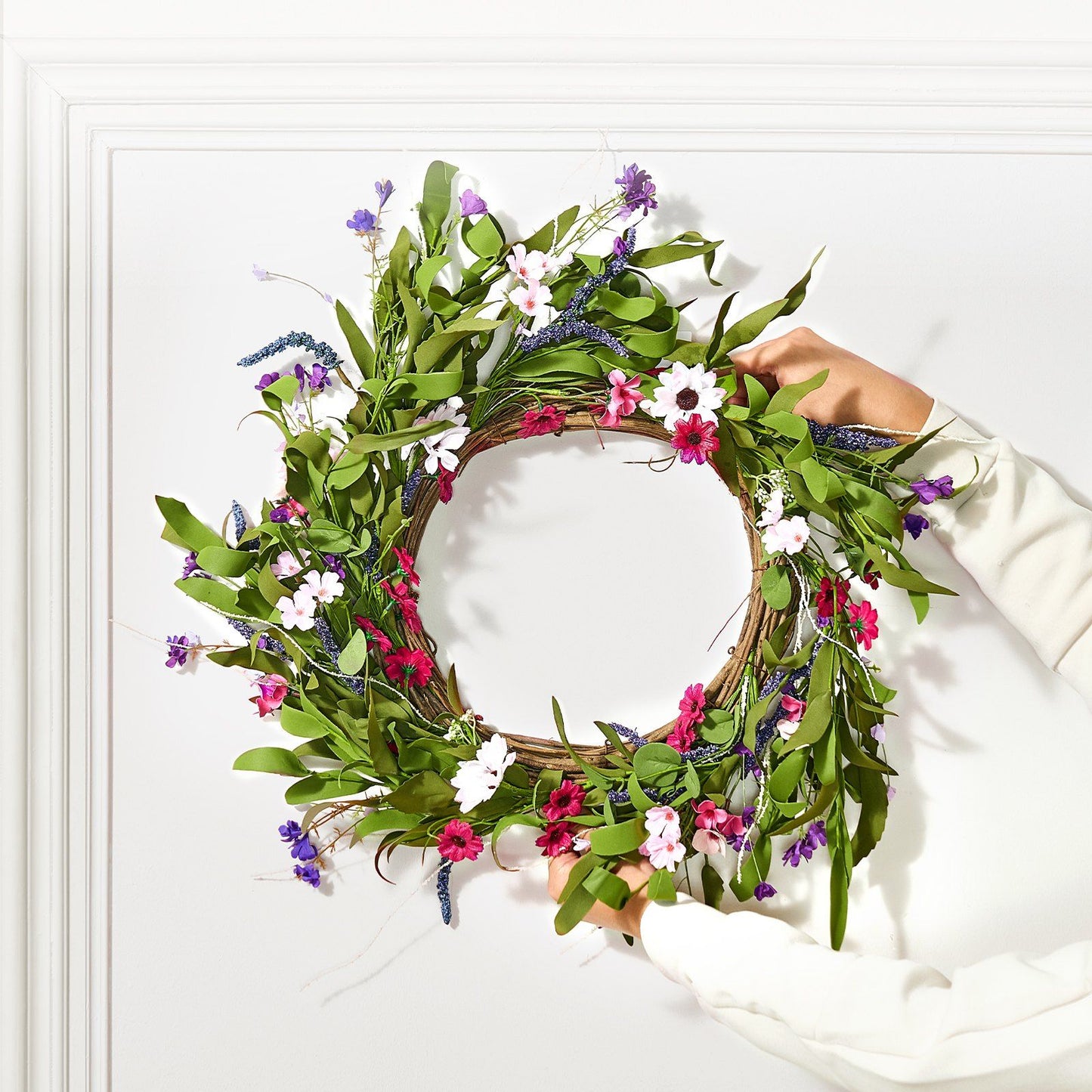 Hands holding an artificial pink daisy wreath with green leaves and purple flowers against a white wall