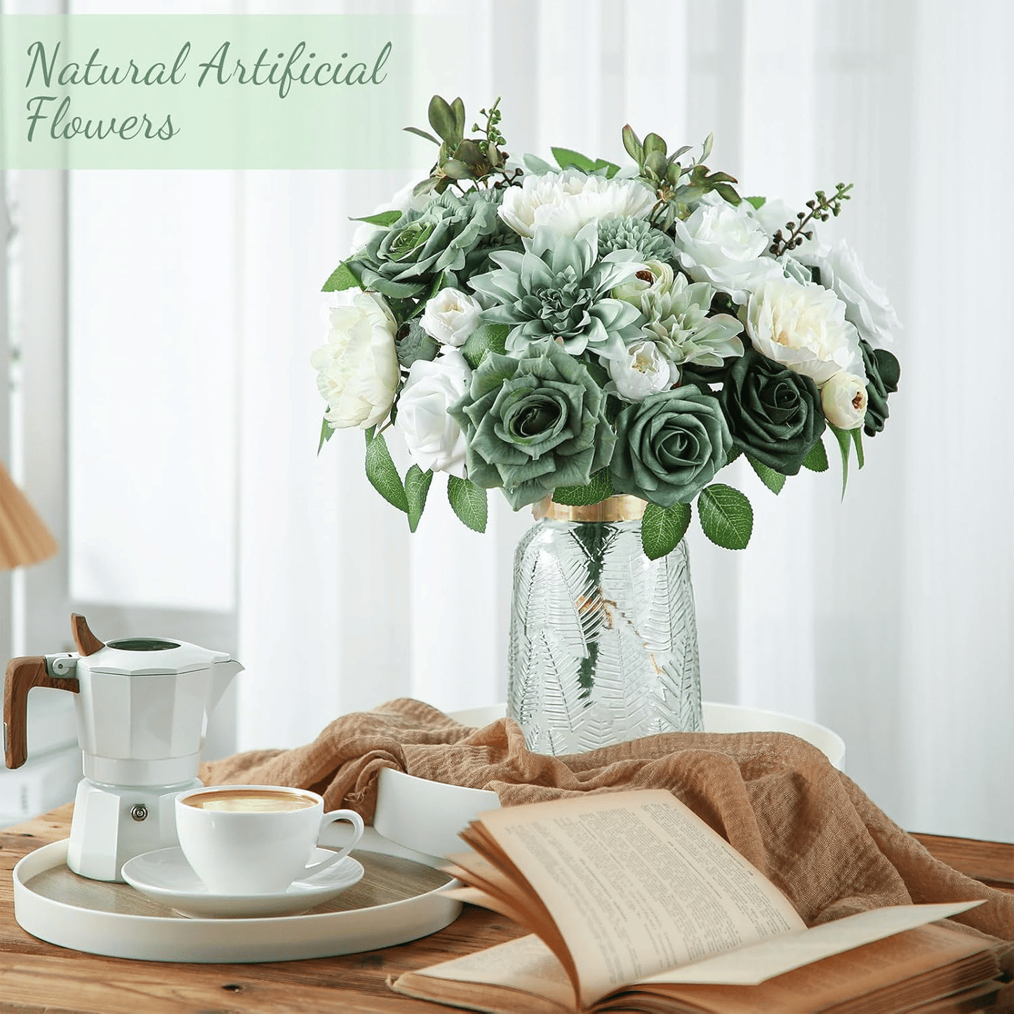 White and green artificial flower arrangement in glass vase on table next to coffee cup and book
