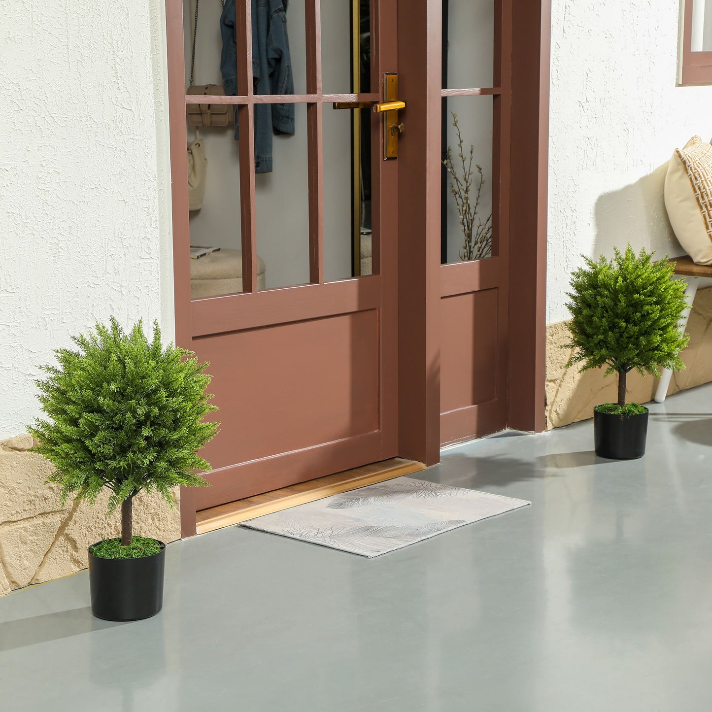 Two green indoor outdoor fake plants in black pots placed on patio near a brown door entrance