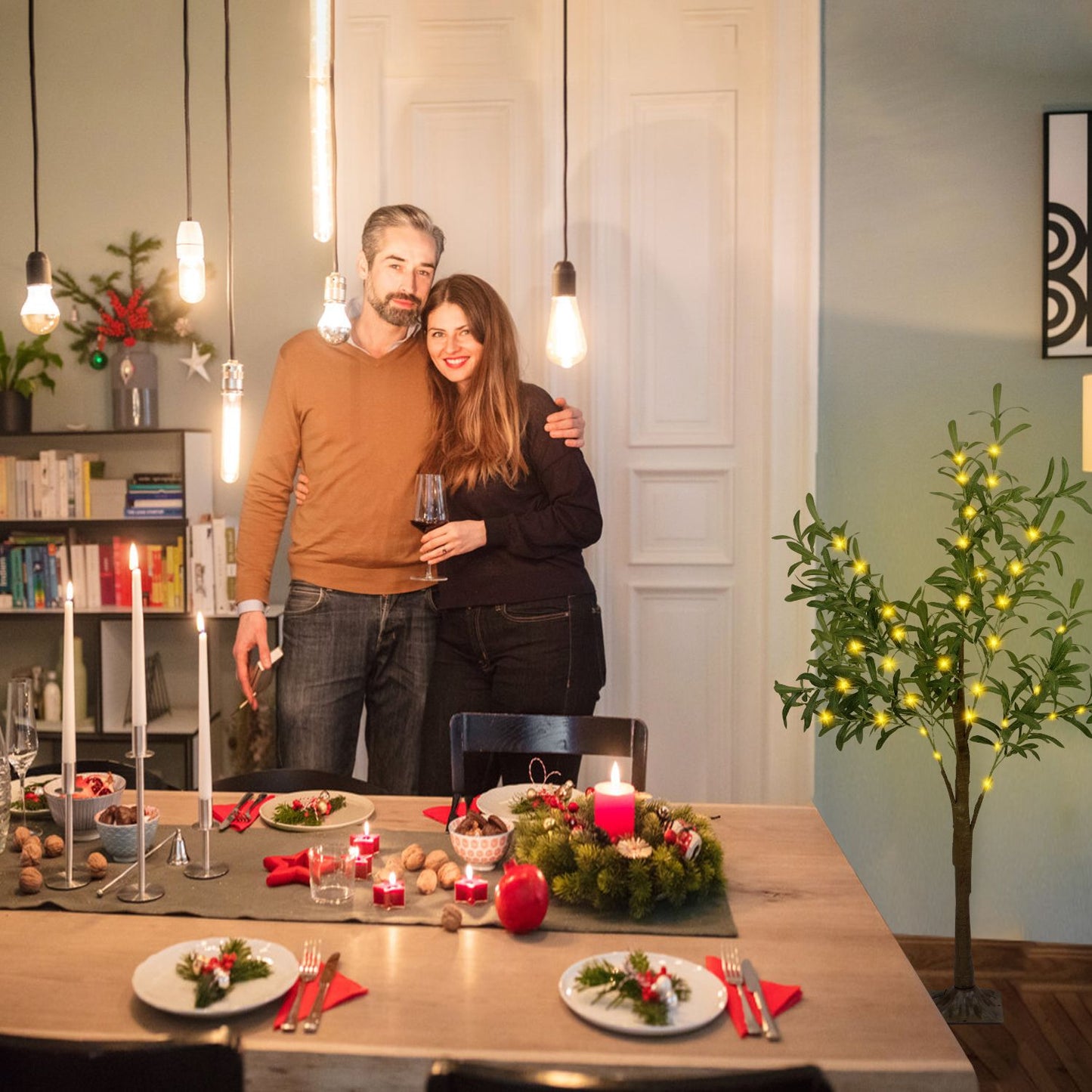 LED olive tree decor lit with warm lights next to dining table set for holiday meal with couple standing behind