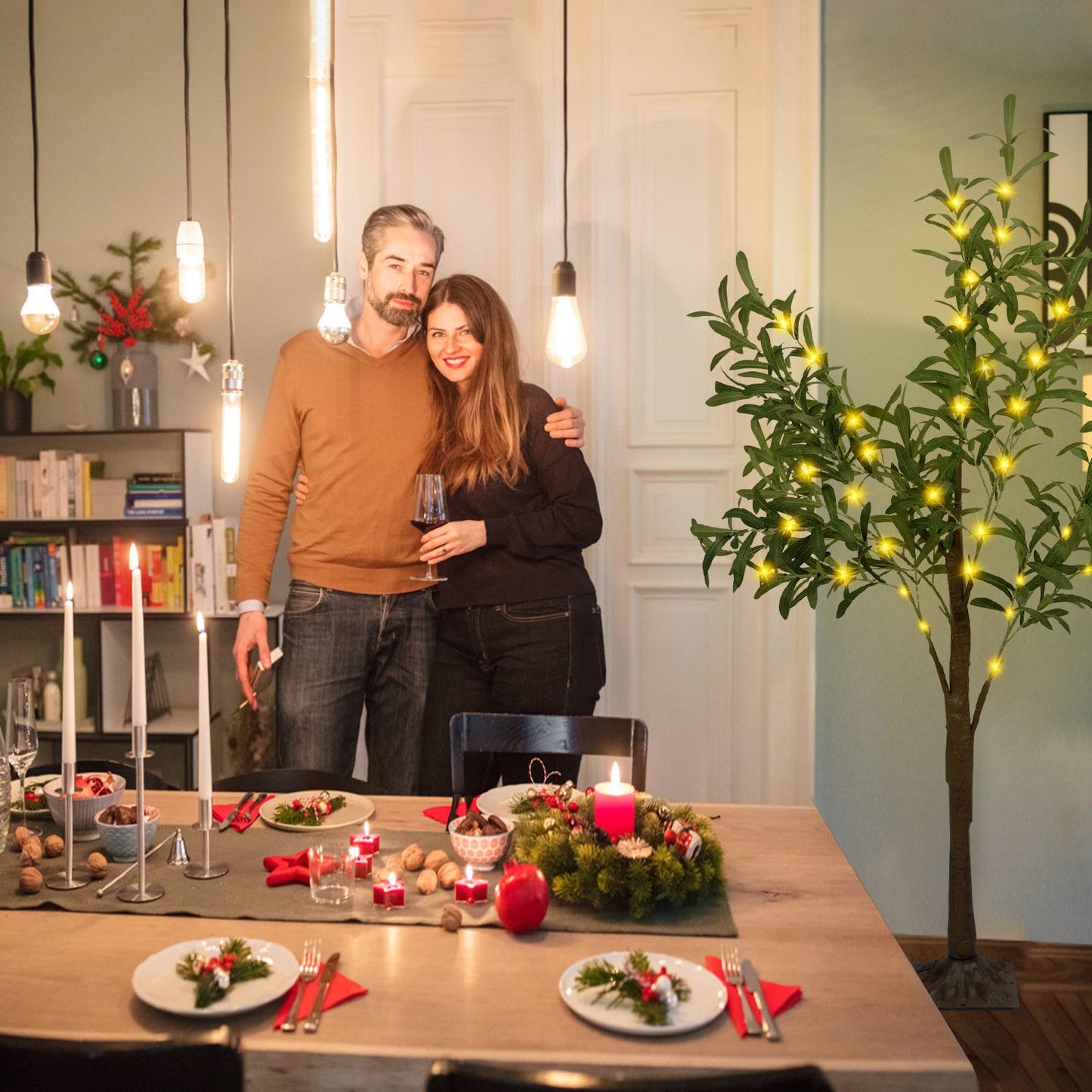 LED olive tree decor with warm lights beside a cozy holiday dining table and a smiling couple indoors