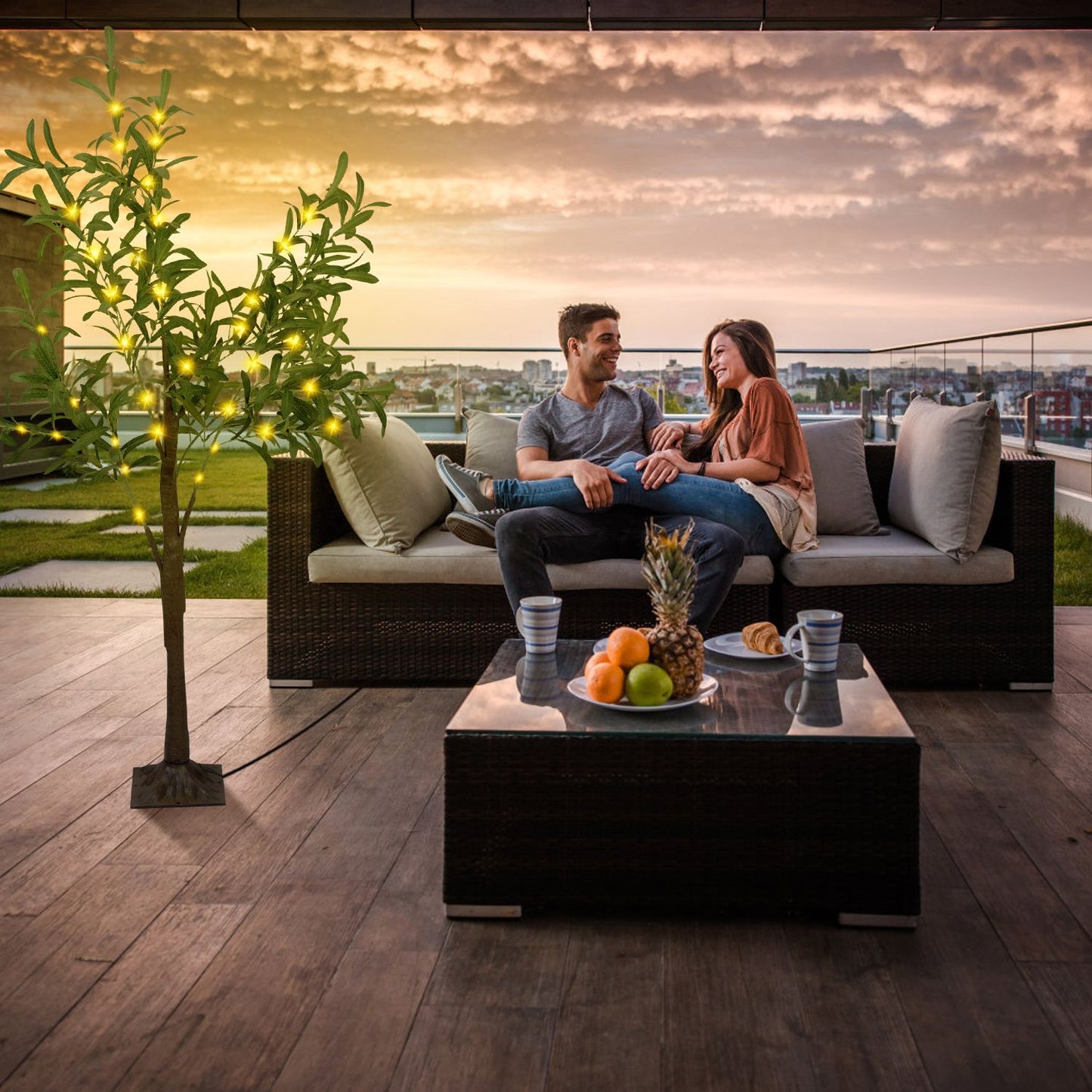 Couple sitting on outdoor sofa next to LED olive tree decor with glowing lights at sunset