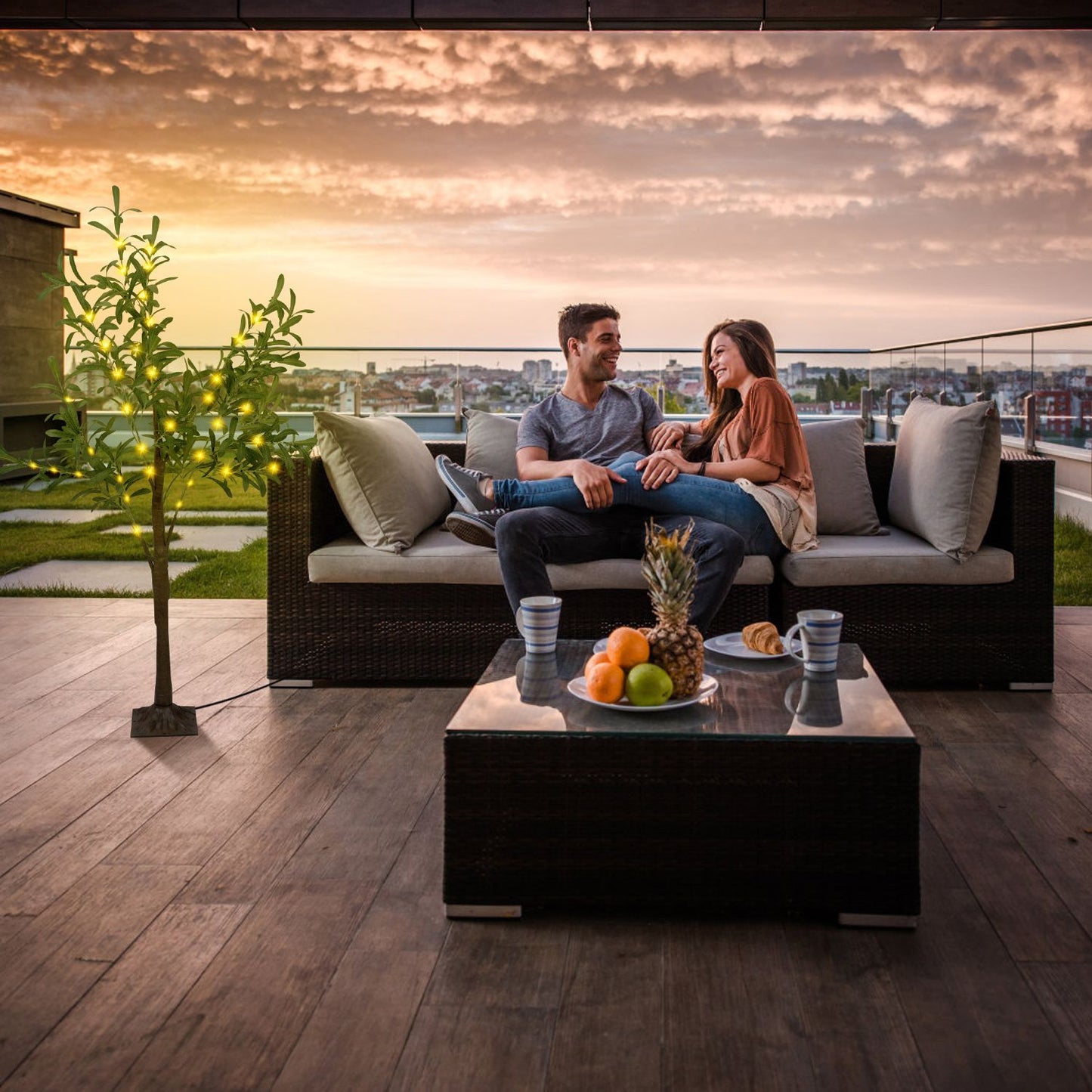 Couple sitting on outdoor sofa with LED olive tree decor lit up at sunset on rooftop patio
