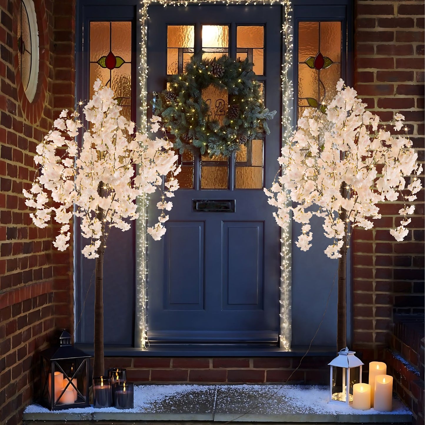 Lighted cherry blossom tree decor on both sides of a blue front door with holiday wreath and candles