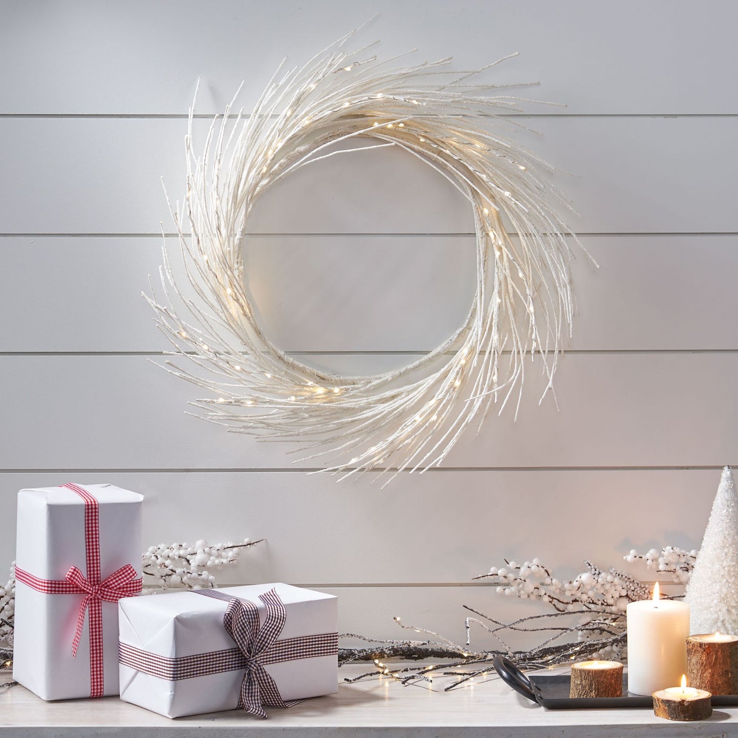 Elegant lighted paper wreath with delicate white branches hanging on a wall above wrapped gifts and candles