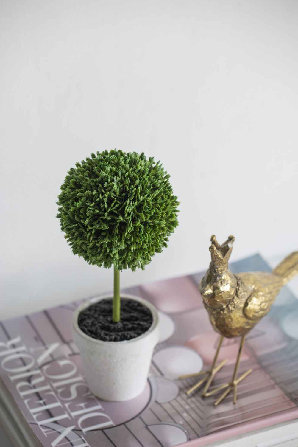Mini faux topiary set in white pot placed on a book next to a gold decorative bird figurine