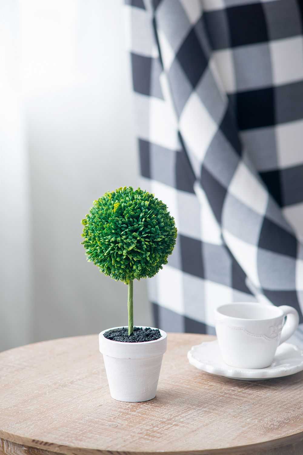 Mini faux topiary set with green round foliage in white pot on wooden table next to white teacup and saucer