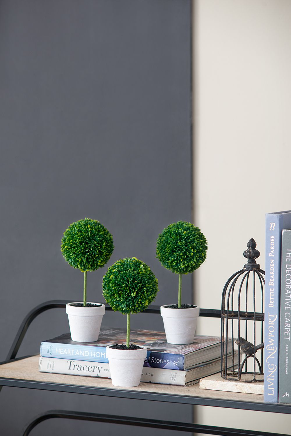 Mini faux topiary set with three green artificial plants in white pots displayed on books on a wooden table