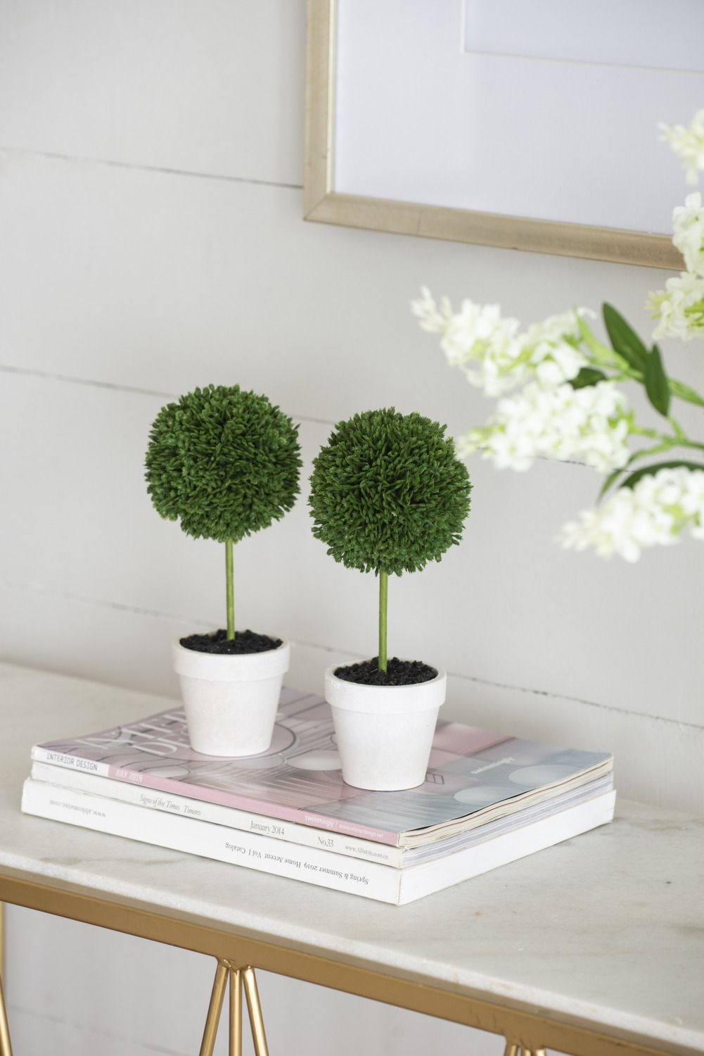 Mini faux topiary set with two green ball-shaped plants in white pots on a stack of books