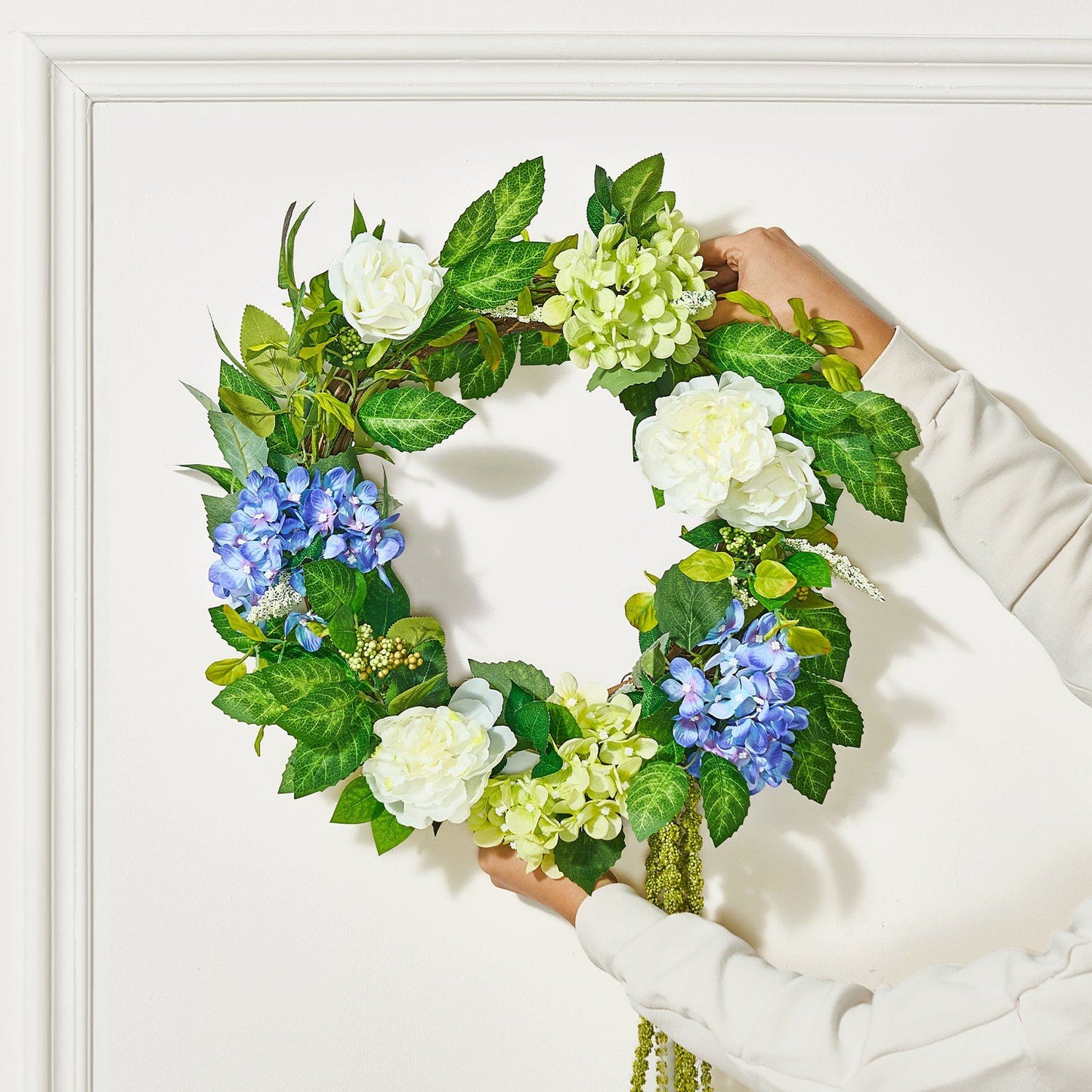 Hands holding an artificial hydrangea wreath with white, blue, and green flowers and lush green leaves