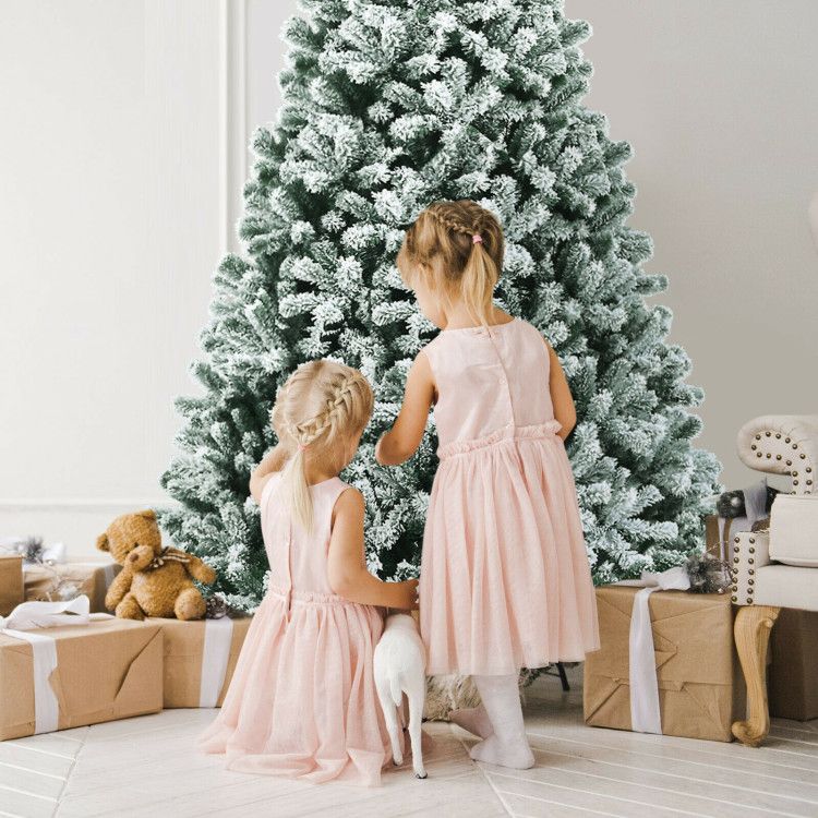 Two girls in pink dresses decorating a pre-lit snow flocked Christmas tree with gifts and teddy bear nearby
