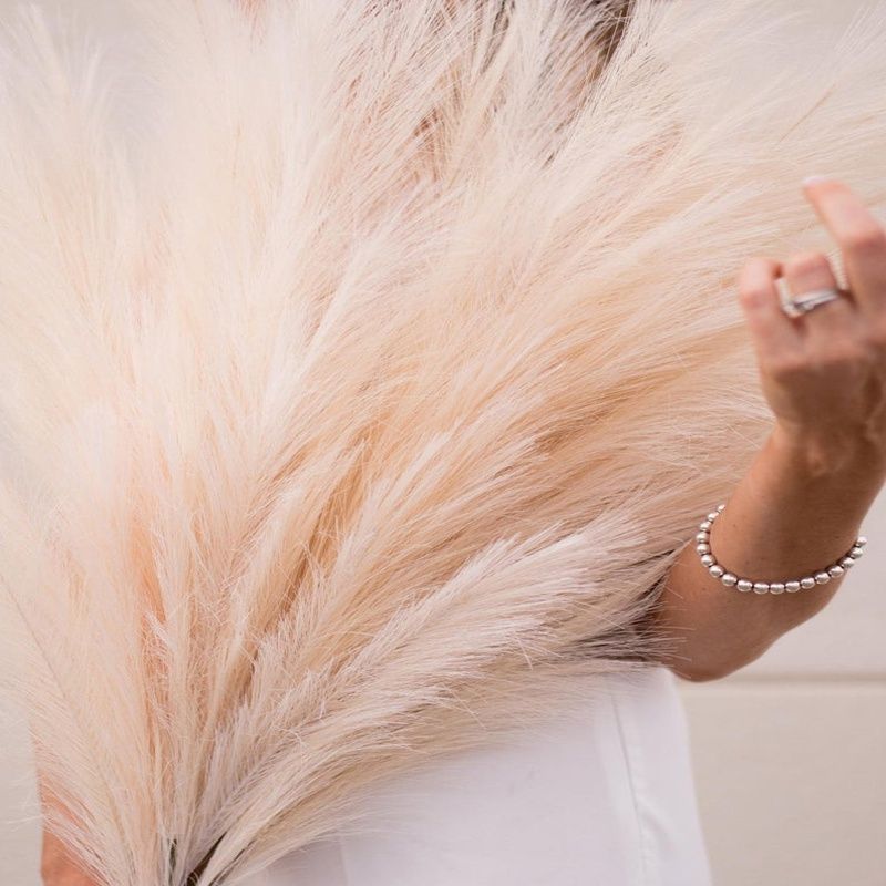 Close-up of tall artificial pampas grass decor held by person wearing white and silver bracelet