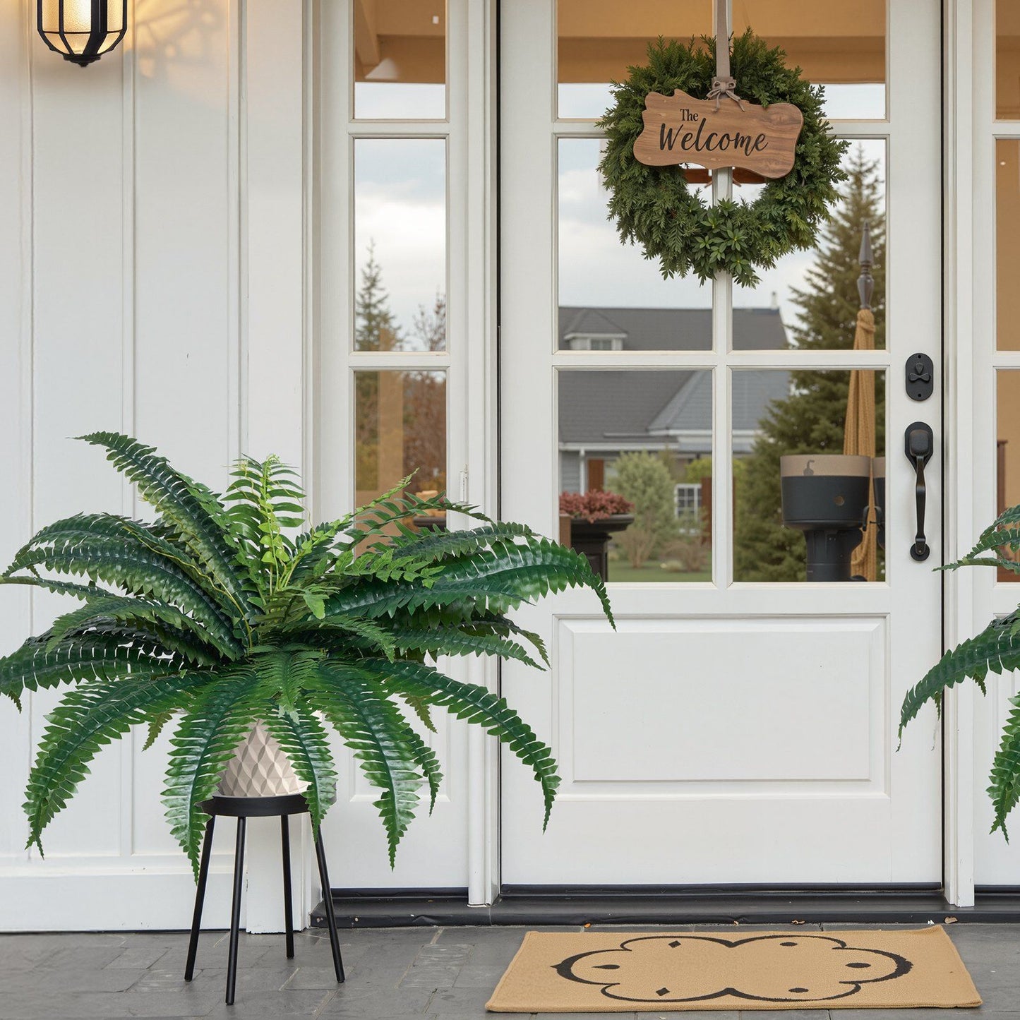 Artificial Boston fern set in white textured pot on black stand by front door with welcome wreath
