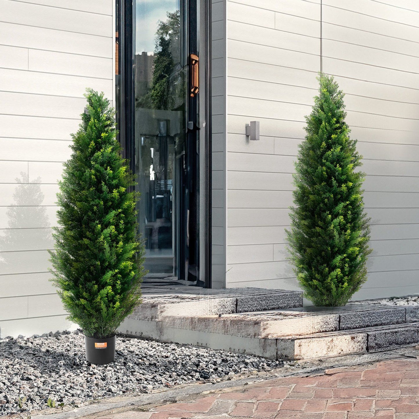 Two realistic faux cedar topiary trees in black pots placed outside a modern home entrance
