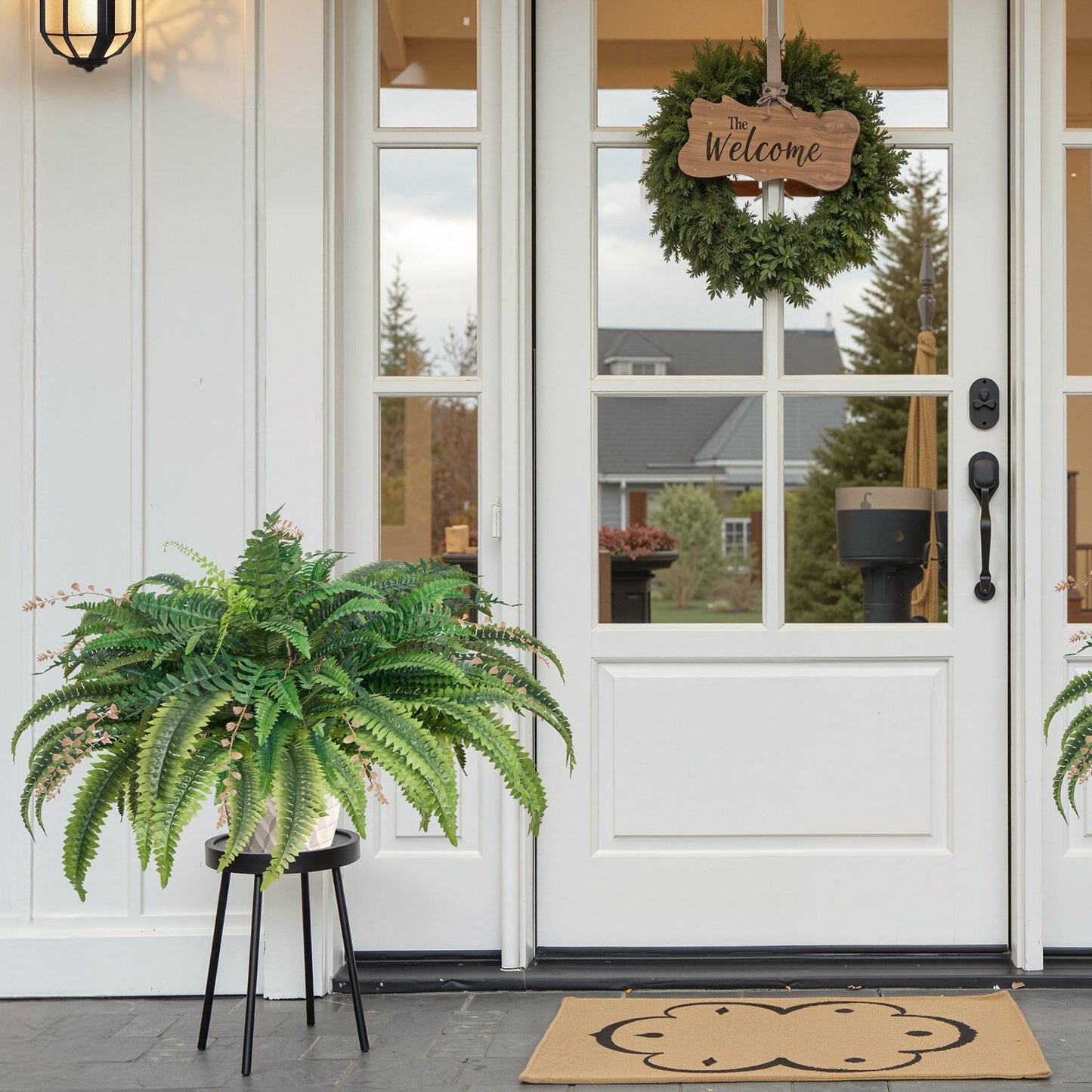 Large artificial Boston fern in a black stand placed on a porch next to a white door with a welcome wreath