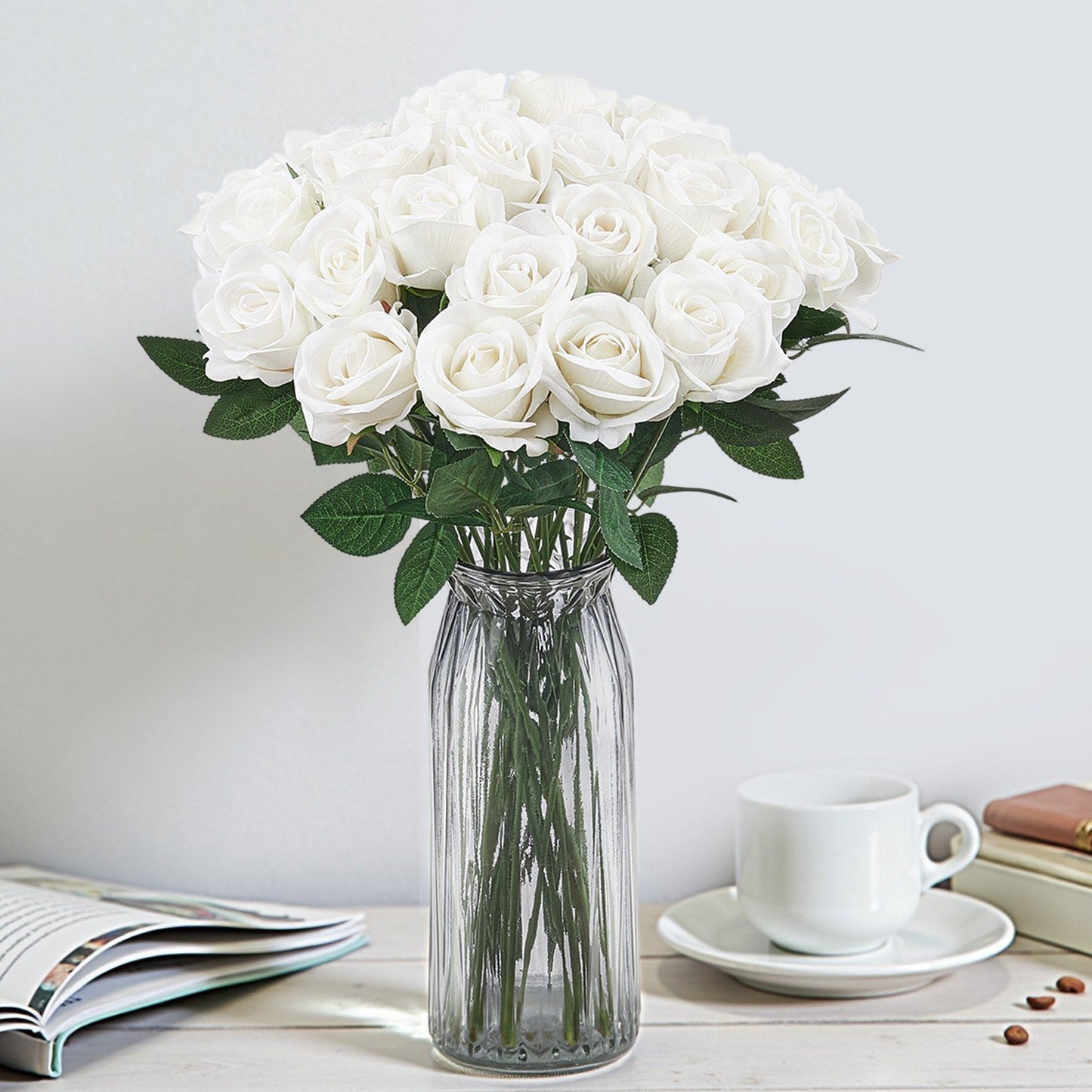 White artificial roses bouquet in clear glass vase on table with cup and open book