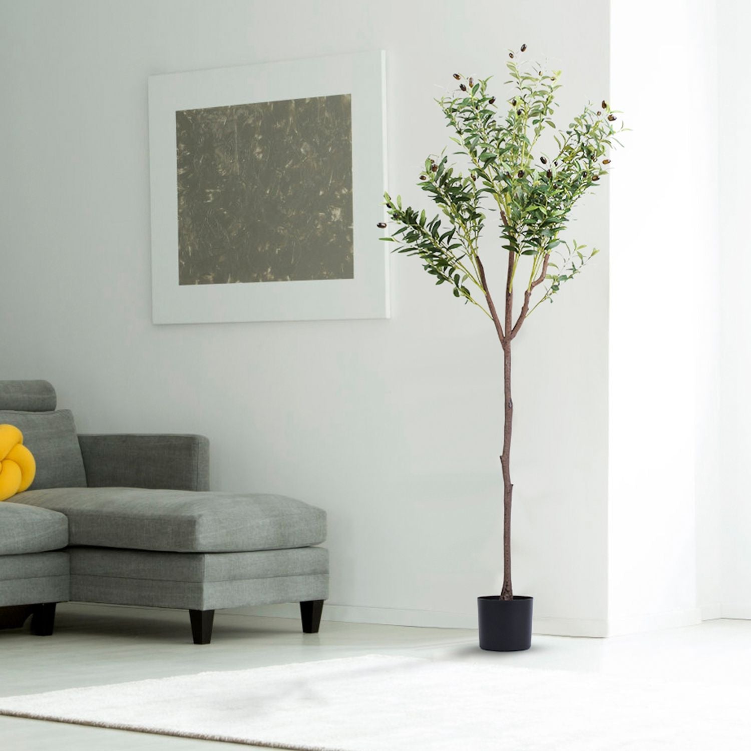Tall artificial olive tree for home decor in a black pot next to a gray sectional sofa in a bright living room
