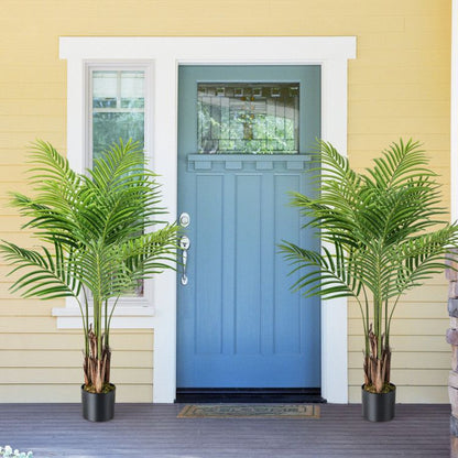 Two artificial palm trees for home decor in black pots placed on a porch with a blue front door