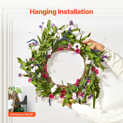 Hands hanging an artificial pink daisy wreath with green leaves and mixed flowers on a white wall