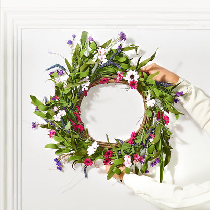 Hands holding an artificial pink daisy wreath with green leaves and purple flowers against a white wall