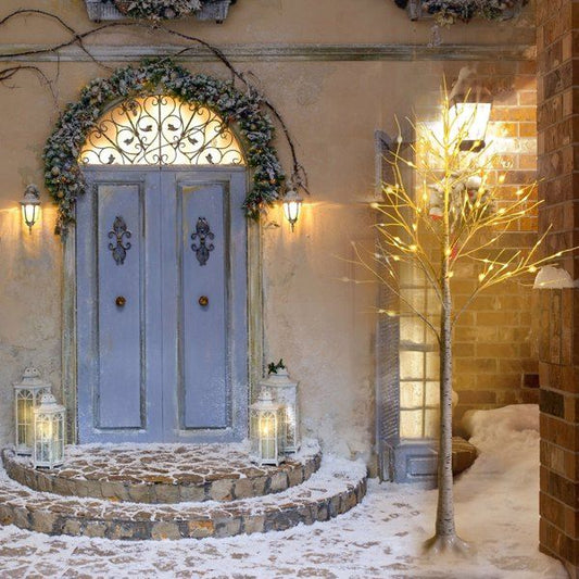 Lighted birch tree decor with warm white lights displayed outside a rustic blue door in a snowy setting