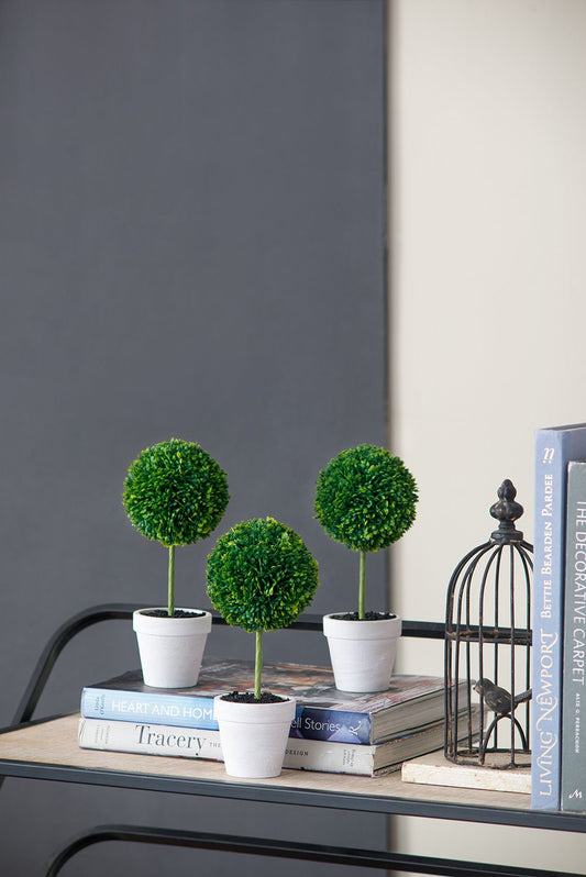 Mini faux topiary set with three green artificial plants in white pots displayed on books on a wooden table