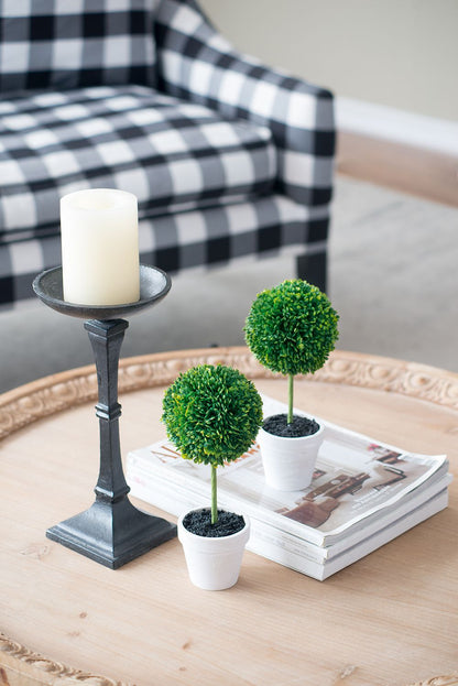 Mini faux topiary set with two small green plants in white pots on a wooden table beside a black candle holder