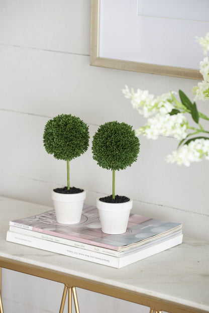 Mini faux topiary set with two green ball-shaped plants in white pots on a stack of books