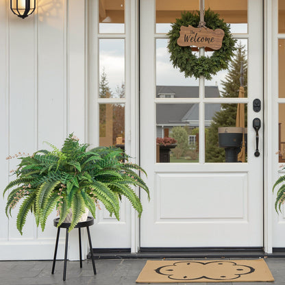 Large artificial Boston fern in a black stand placed on a porch next to a white door with a welcome wreath